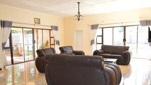 A spacious living room with a large brown leather sofa, a smaller sofa, and a coffee table. There are large windows with curtains, a chandelier, and a door leading outside.
