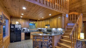 A wooden cabin interior with a kitchen featuring a central island, wooden cabinets, and a staircase. - Mountain View, Hot Tub, Large Deck, Fire Pit Area (Morganton)