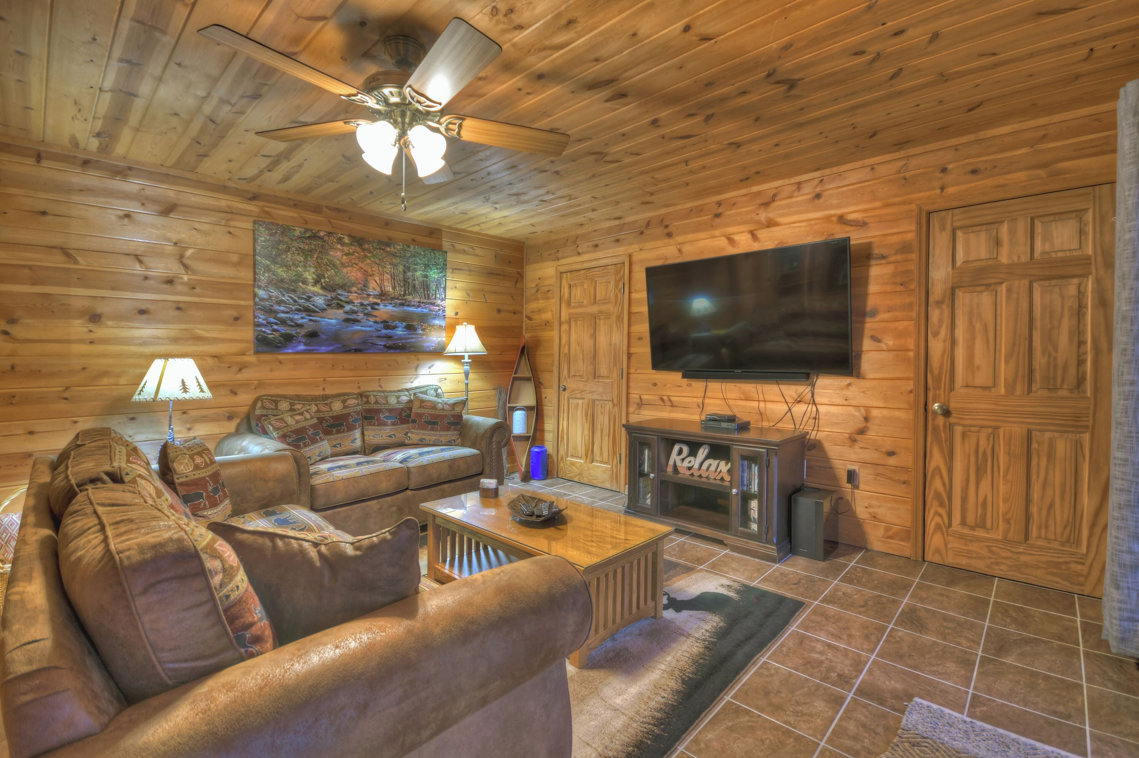 A cozy living room with a wooden interior, a ceiling fan, a flat-screen TV, a sofa, a coffee table, and a cabinet with a 'Relax' sign.
