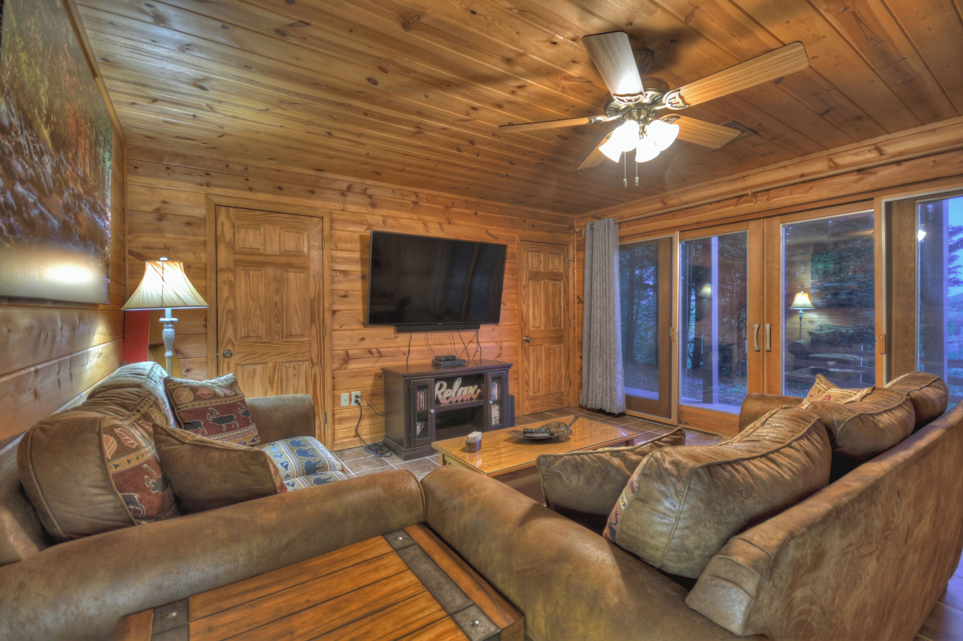 A log cabin interior with a wooden ceiling, a ceiling fan, a flat-screen TV mounted on the wall, a leather sofa with patterned cushions, a coffee table, and a sliding glass door leading outside.