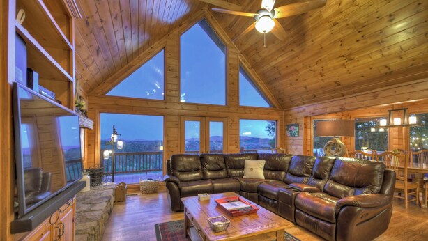A spacious living room with a large leather sectional sofa, wooden flooring, and a view of the outdoors through large windows. - Mountain View, Hot Tub, Large Deck, Fire Pit Area (Morganton)