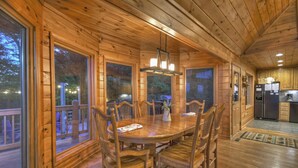 A wooden cabin interior with a dining table, chairs, a chandelier, and a kitchen area. - Mountain View, Hot Tub, Large Deck, Fire Pit Area (Morganton)