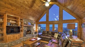 A cozy cabin interior with a stone fireplace, leather sofa, wooden shelves, and a large TV. - Mountain View, Hot Tub, Large Deck, Fire Pit Area (Morganton)