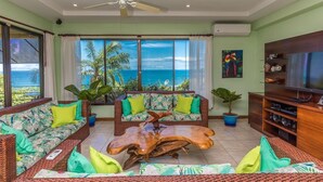 A spacious living room with a large window overlooking the ocean, a ceiling fan, and a wooden coffee table. - Vista Oceana, Stunning Ocean View Villa (Quepos)