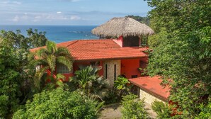 A red-roofed building with a thatched roof structure, surrounded by lush greenery and overlooking a body of water. - Vista Oceana, Stunning Ocean View Villa (Quepos)