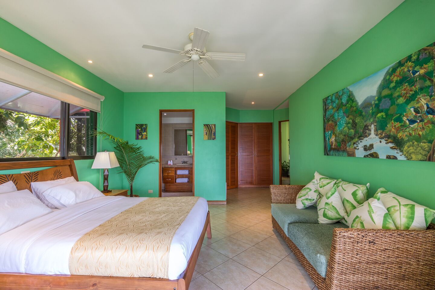 A bedroom with a bed, a sofa, a ceiling fan, and a painting of a jungle scene.