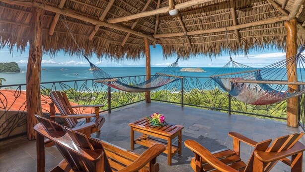 A thatched-roof outdoor seating area with wooden chairs and a table, overlooking a coastal view. - Vista Oceana, Stunning Ocean View Villa (Quepos)