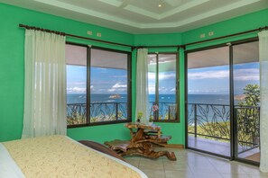 A room with a bed, a wooden side table, and large windows offering ocean views. - Vista Oceana, Stunning Ocean View Villa (Quepos)