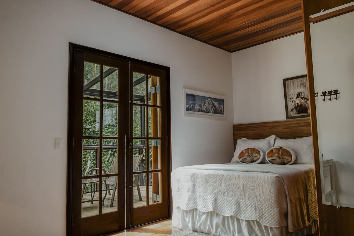 Un dormitorio con techo de madera, una cama con ropa blanca, un amplio ventanal corredero que da a un espacio exterior con sillas y un cuadro en la pared.