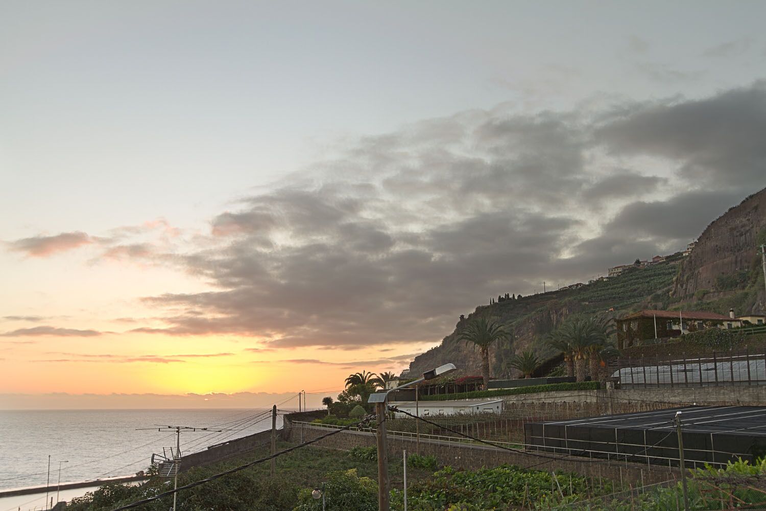 A coastal landscape with a sunset, a sea wall, and a building with a sloped roof.