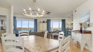 Un comedor amplio con una mesa de madera y sillas blancas, un candelabro y vistas al mar a través de amplias ventanas.
