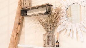 Une étagère murale sur laquelle repose un panier tressé contenant des plantes séchées, un petit flacon et un miroir décoratif.