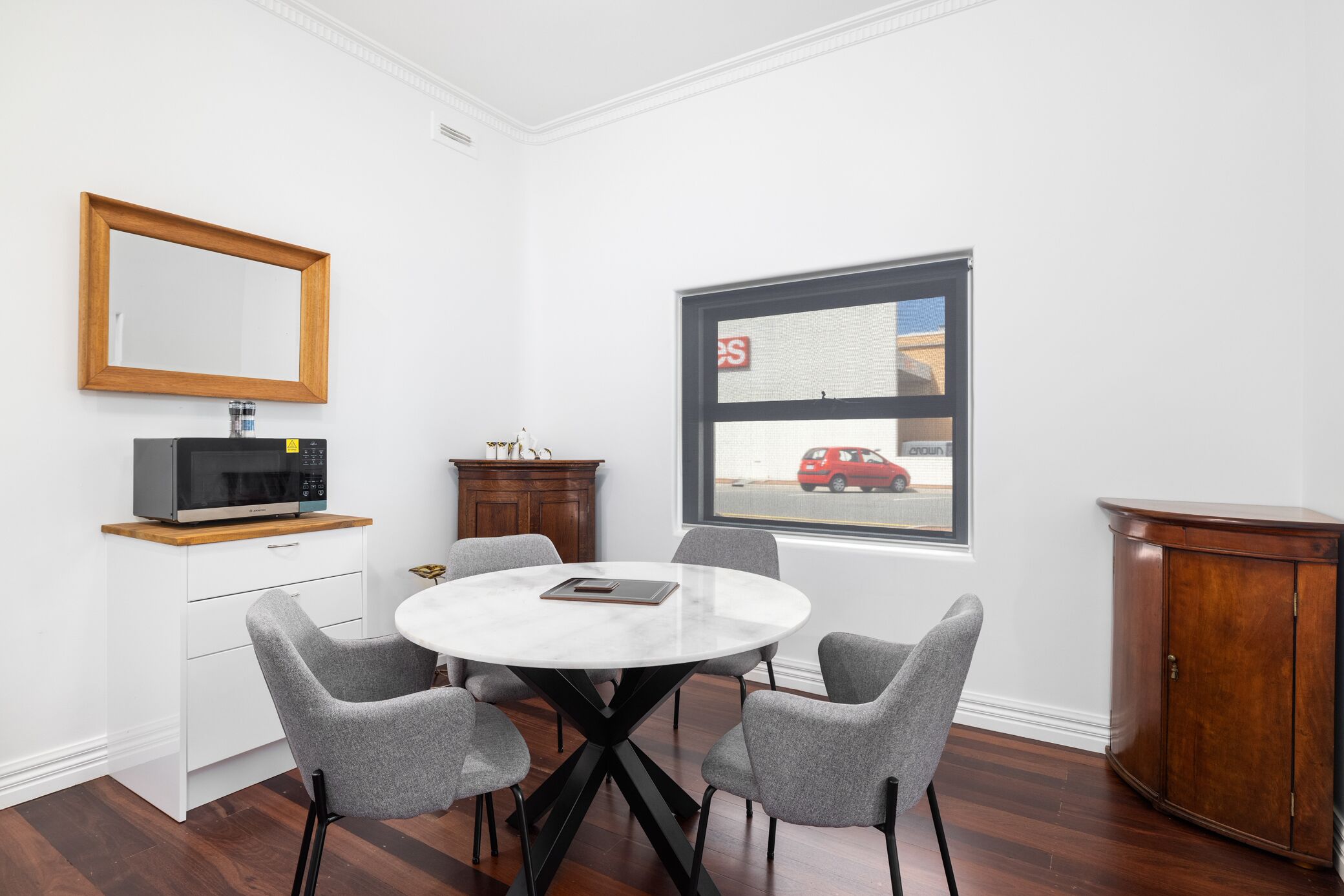 A modern room with a round table, four grey chairs, a wooden cabinet, a microwave, and a large window with a view of a red car.