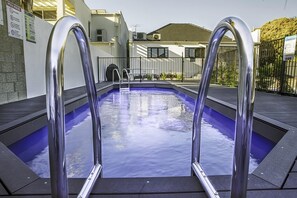 Piscina com escadas de metal, cercada por uma grade e um edifício ao fundo.