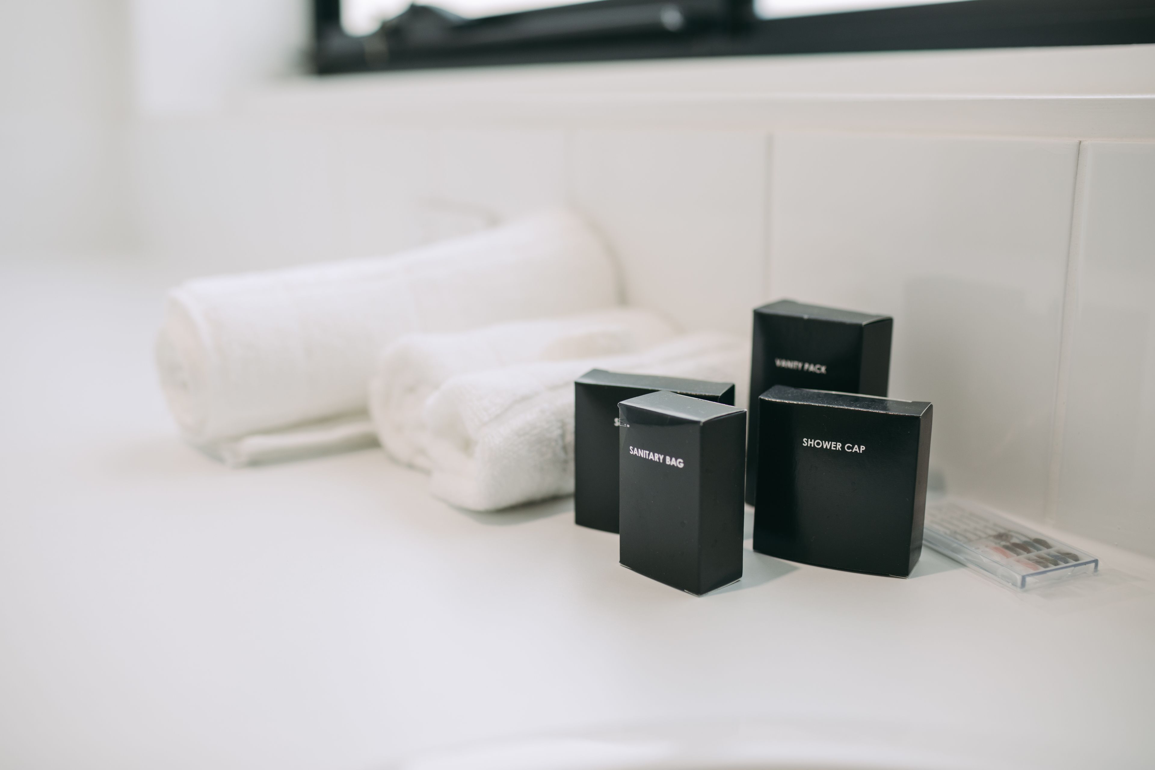 A bathroom with a white sink, two rolled towels, and three black boxes labeled "SANITARY BAG," "SHOWER CAP," and "SHOWER CAP."