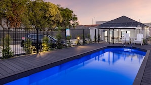A swimming pool with a wooden deck, surrounded by a black fence. There are white chairs and a table near the pool, and a building with a dark roof in the background.