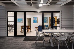 Un comedor moderno con una mesa y sillas blancas, un ventilador de techo y vistas a través de puertas de vidrio hacia otro espacio con sillas y mesas.