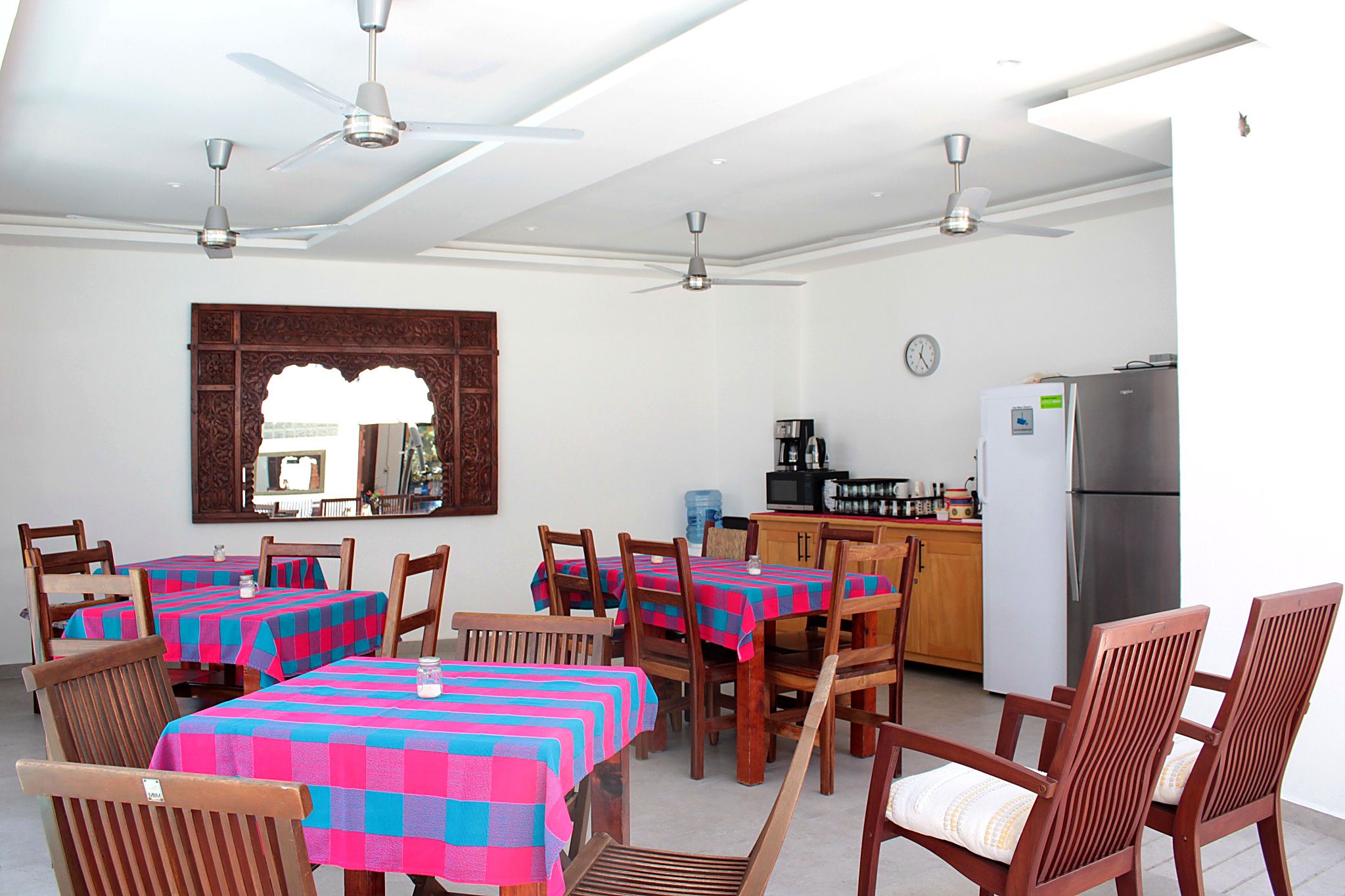 Un comedor con mesas dispuestas para cuatro personas, una zona de cocina con refrigerador y un espejo grande en la pared.