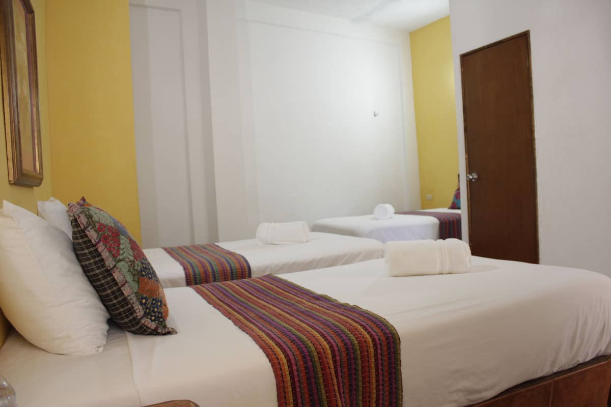 Quarto de hotel com duas camas, cada uma com roupa de cama branca e almofadas coloridas. Há uma porta de madeira e uma parede amarela.