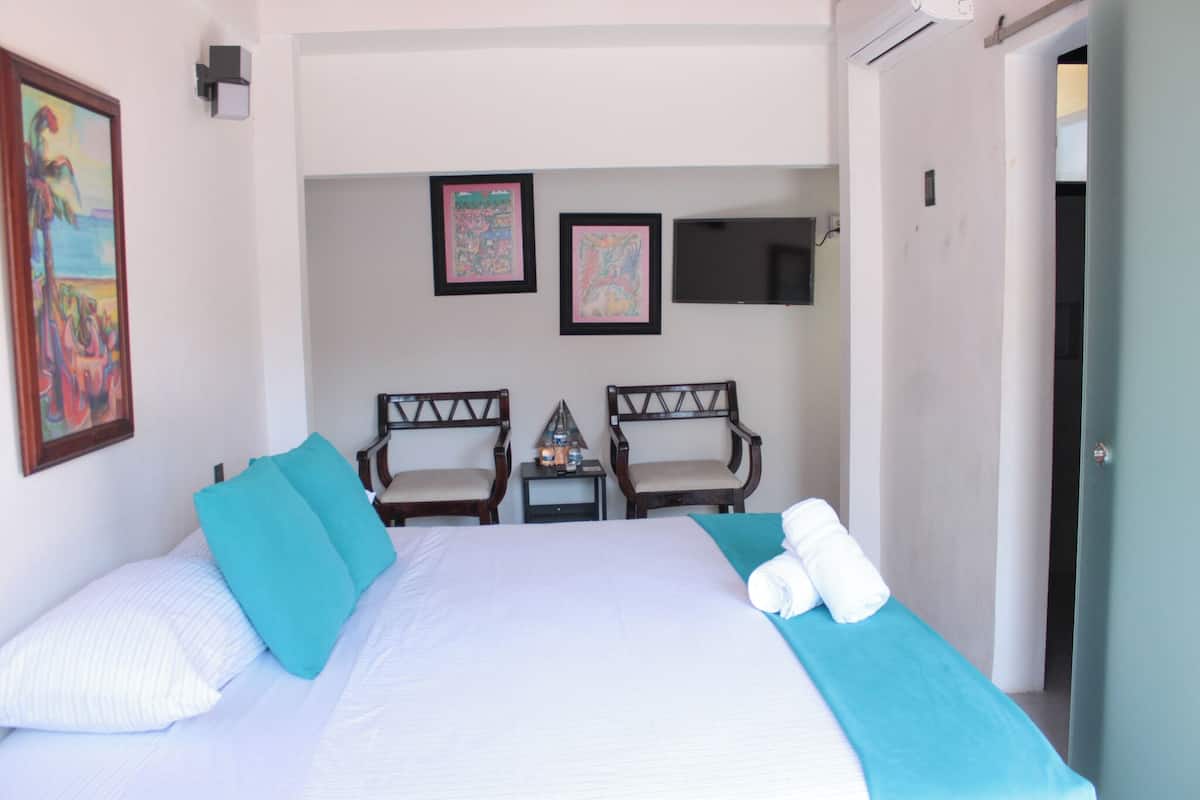 Quarto de hotel com uma cama grande, duas cadeiras, uma mesinha com uma garrafa e copos, e uma TV de tela plana fixada na parede.