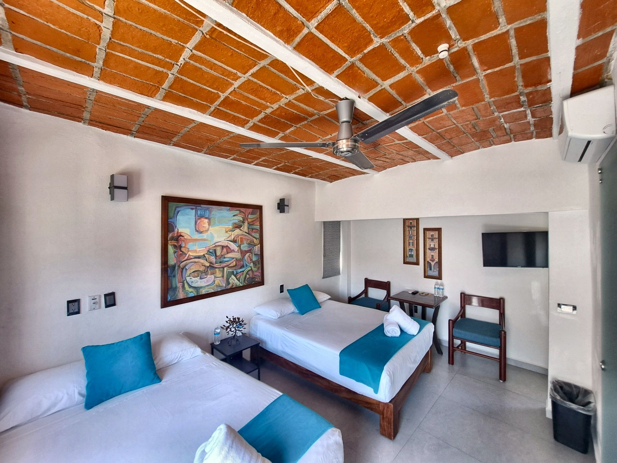 Habitación de hotel con dos camas, un ventilador de techo, un cuadro, una mesita y un bote de basura.