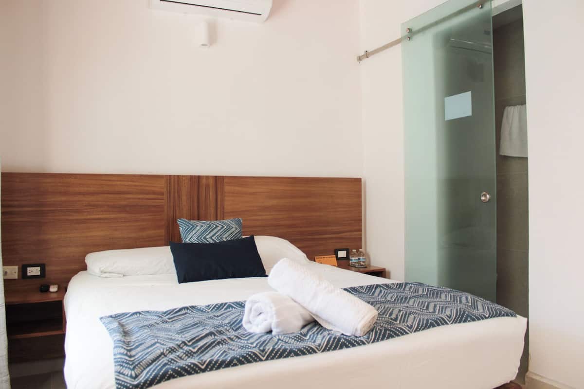 Cama bem arrumada com colcha estampada, toalha enrolada e mesa de cabeceira com uma garrafa e um pequeno objeto.