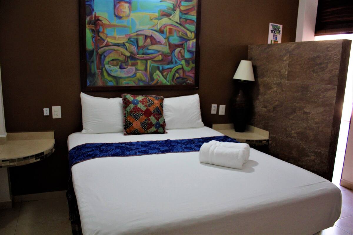 Quarto de hotel com uma cama grande, um quadro abstrato colorido na parede, uma mesa de cabeceira com abajur e uma parede de detalhes em pedra.