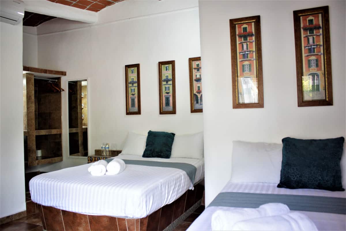 Quarto de hotel com duas camas, banheiro com chuveiro e corredor com quadros nas paredes.