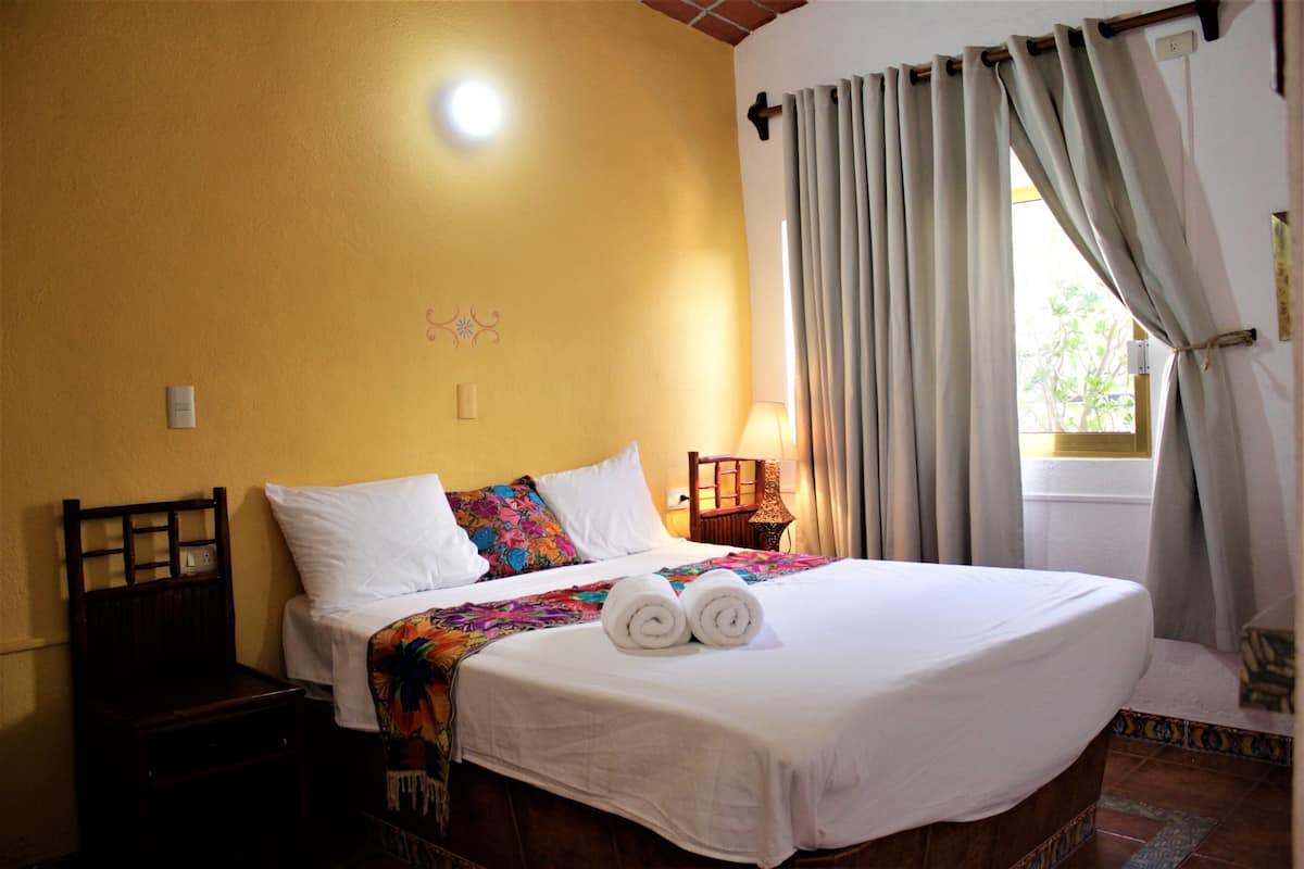 Uma cama bem arrumada com roupa de cama branca e toalhas dobradas, uma mesinha de cabeceira de madeira, um abajur e uma janela com cortinas.