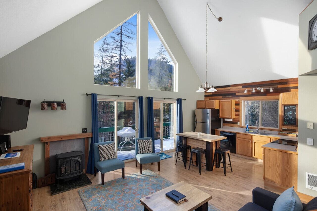 A spacious living area with a fireplace, seating, and a kitchen with wooden cabinets and large windows.