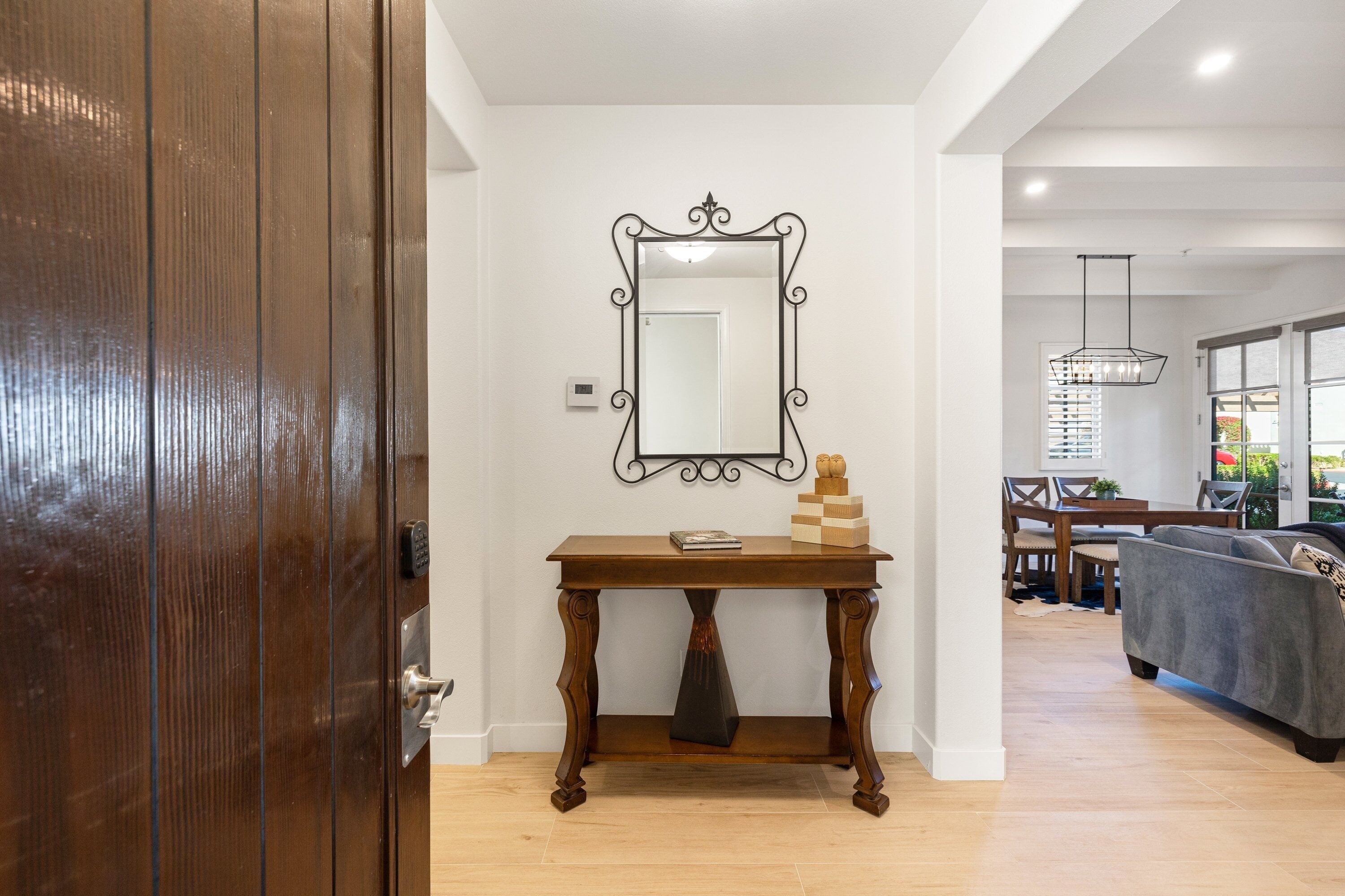 A wooden door, a mirror, a small table with a book and a chess set, a gray sofa, and a dining area with chairs and a hanging light fixture.