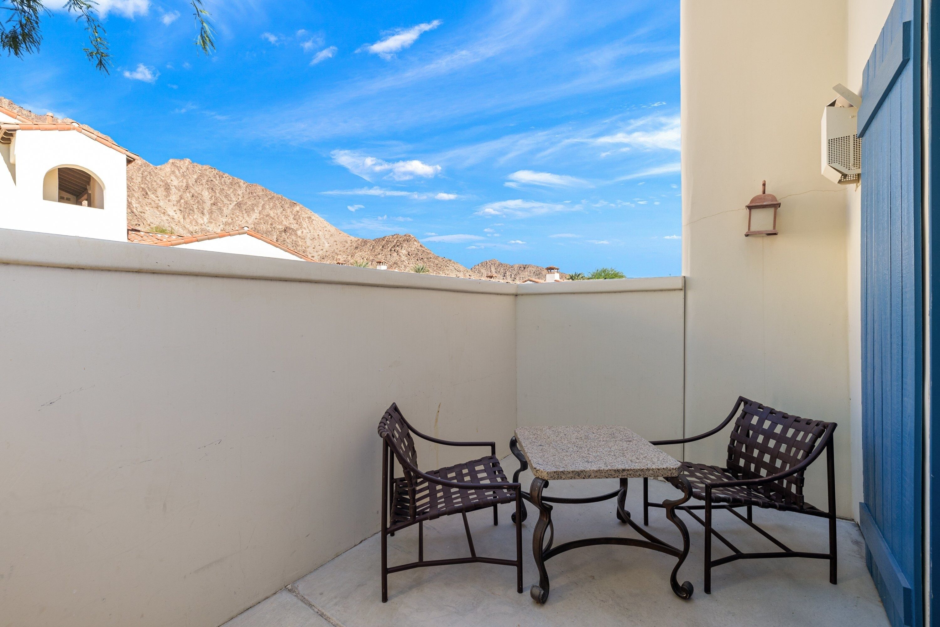 A patio with two metal chairs and a table, a blue door, and a white building with a mountain backdrop.
