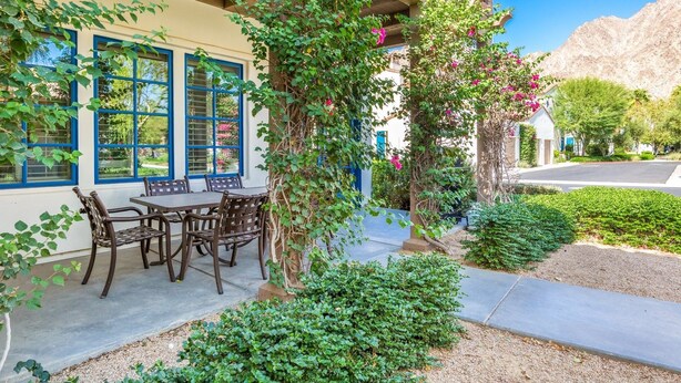 Eine Terrasse mit Tisch und Stühlen, umgeben von Grünpflanzen und einem Gebäude mit blauen Fensterrahmen.