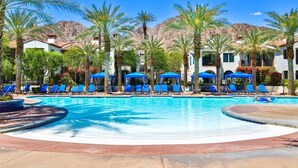 Ein Poolbereich mit blauen Liegestühlen und Sonnenschirmen, umgeben von Palmen und Gebäuden vor einer Bergkulisse.