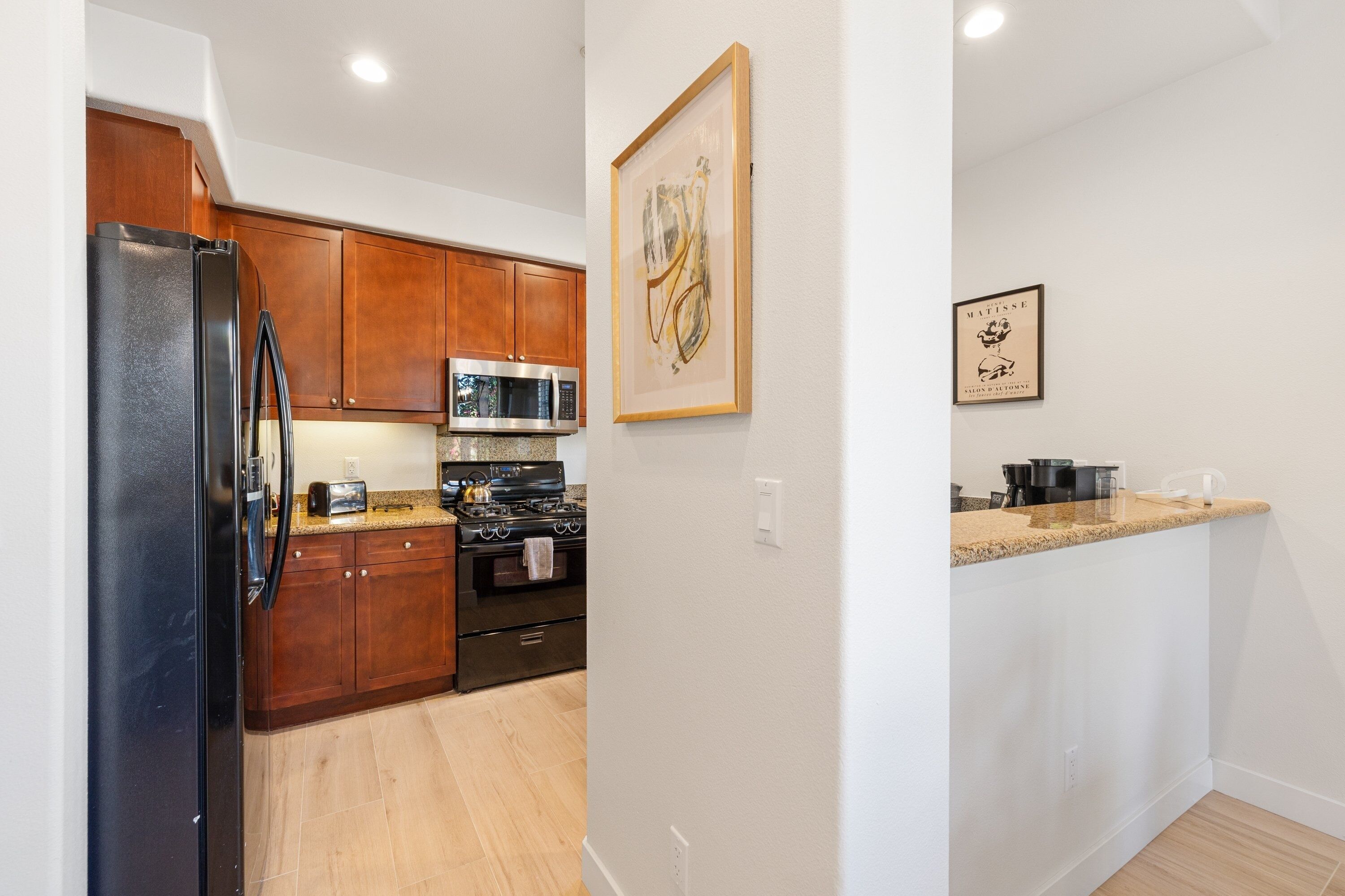 A modern kitchen with wooden cabinets, a stainless steel refrigerator, a microwave, and an oven. There are two framed artworks on the wall.