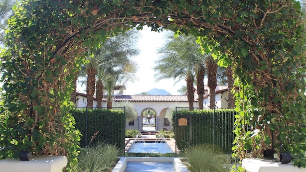 An arched entrance with a pathway leading to a pool, surrounded by lush greenery and palm trees.