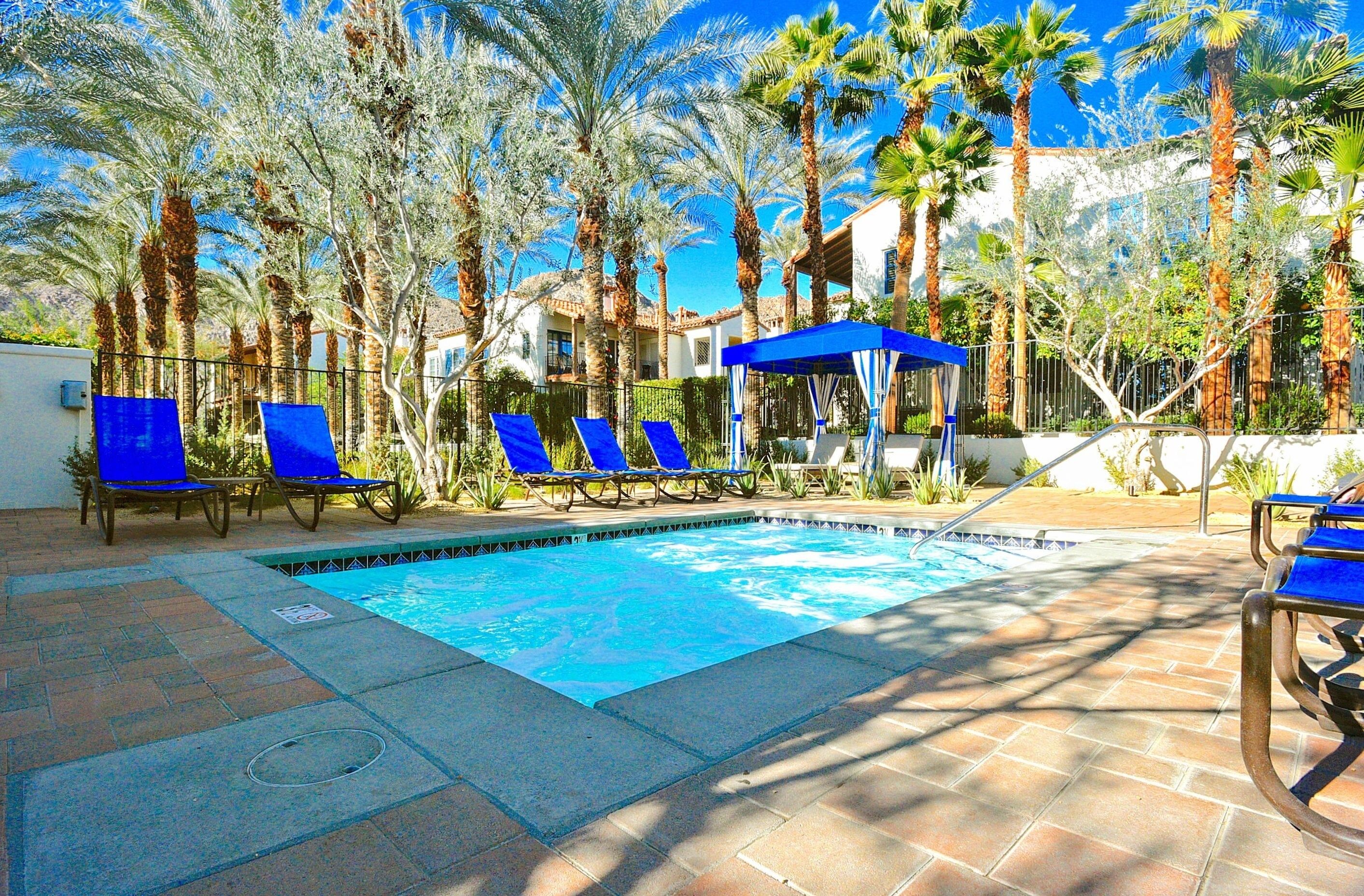 A swimming pool with blue lounge chairs and a covered seating area, surrounded by palm trees and a building in the background.