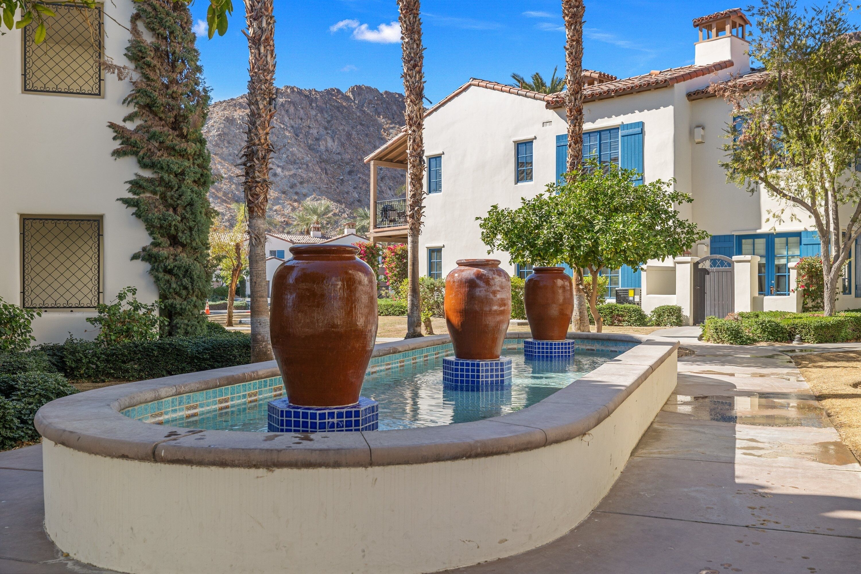 A pool area with large terracotta pots, surrounded by a white building with blue shutters and a mountain backdrop.