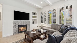 A living room with a fireplace, a sofa, a TV, and large windows with blinds.