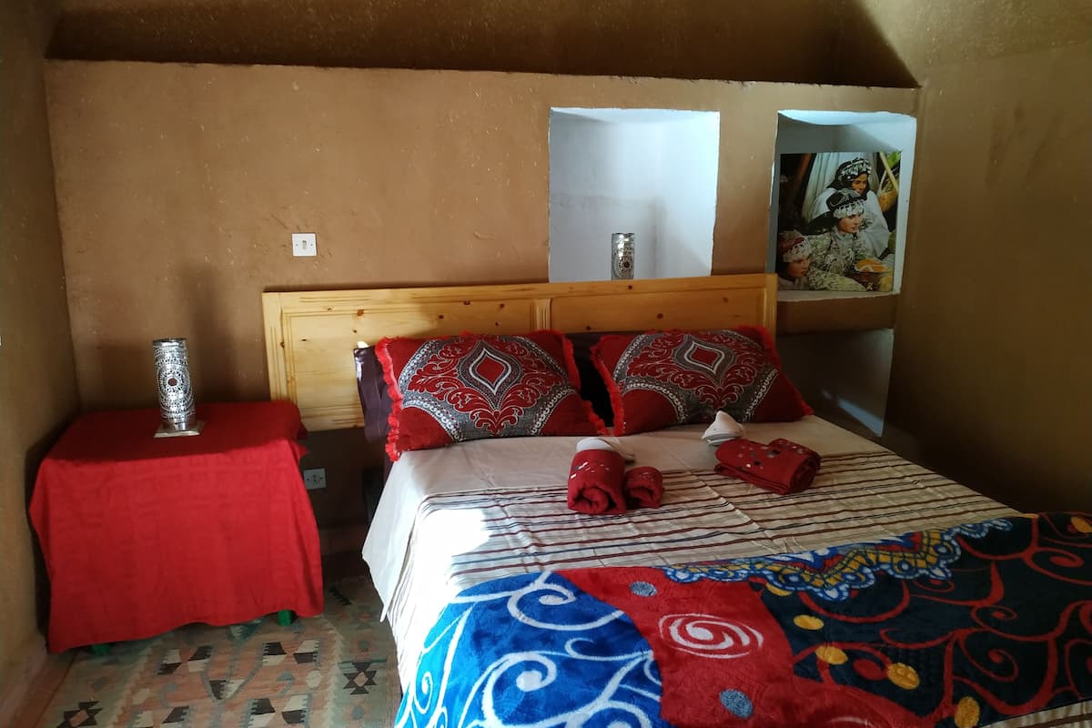 A bedroom with a wooden headboard, a red table with a decorative item, a patterned bedspread, and a colorful blanket.