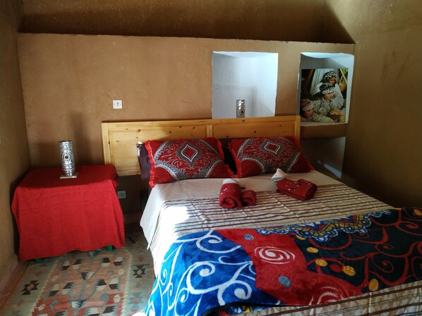 A bedroom with a wooden headboard, a red table with a decorative item, a patterned bedspread, and a colorful blanket. - Riad Dar Zaouia (Zagora)
