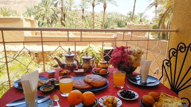 A table set with breakfast items, including pastries, fruits, and beverages, overlooking a scenic view with palm trees and a mountain in the background. - Riad Dar Zaouia (Zagora)