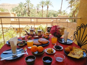 A table set with breakfast items, including pastries, fruits, and beverages, overlooking a scenic view with palm trees and a mountain in the background. - Riad Dar Zaouia (Zagora)