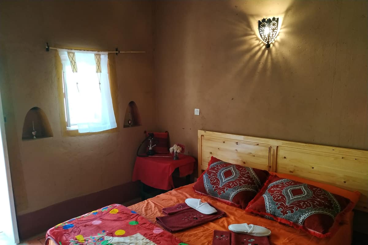 A bedroom with a wooden bed, colorful bedding, a small table with a hookah, a wall-mounted light fixture, and a window with curtains.