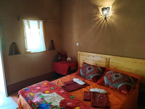 A bedroom with a wooden bed, colorful bedding, a small table with a hookah, a wall-mounted light fixture, and a window with curtains. - Riad Dar Zaouia (Zagora)