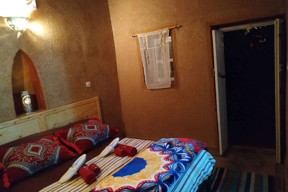 A bedroom with a bed, pillows, a nightstand, a mirror, a window with curtains, and a door.