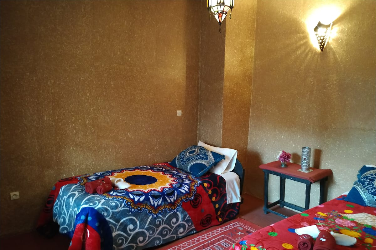 A room with two beds, each with patterned bedding, a small table with a vase, and wall-mounted lights.