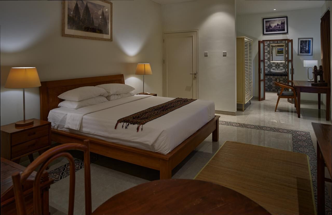 A hotel room with a wooden bed, two bedside lamps, a wooden desk, a chair, and a small table.