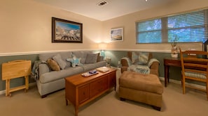 A living room with a sofa, a coffee table, a chair, and a small table with a lamp. There are two framed pictures on the wall.