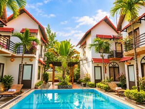 Un área de piscina con una piscina azul cristalina, rodeada por edificios de dos pisos con balcones, plantas en macetas y mobiliario exterior.